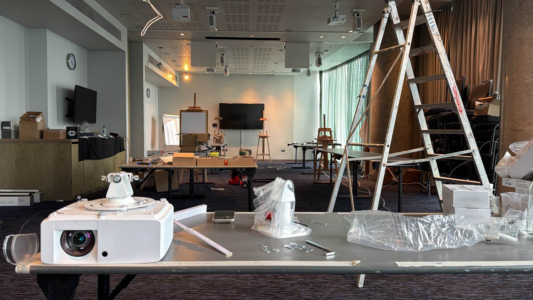 Fit-out day in a function room: a projector with its ceiling mount on a work table, a ladder and easels behind
