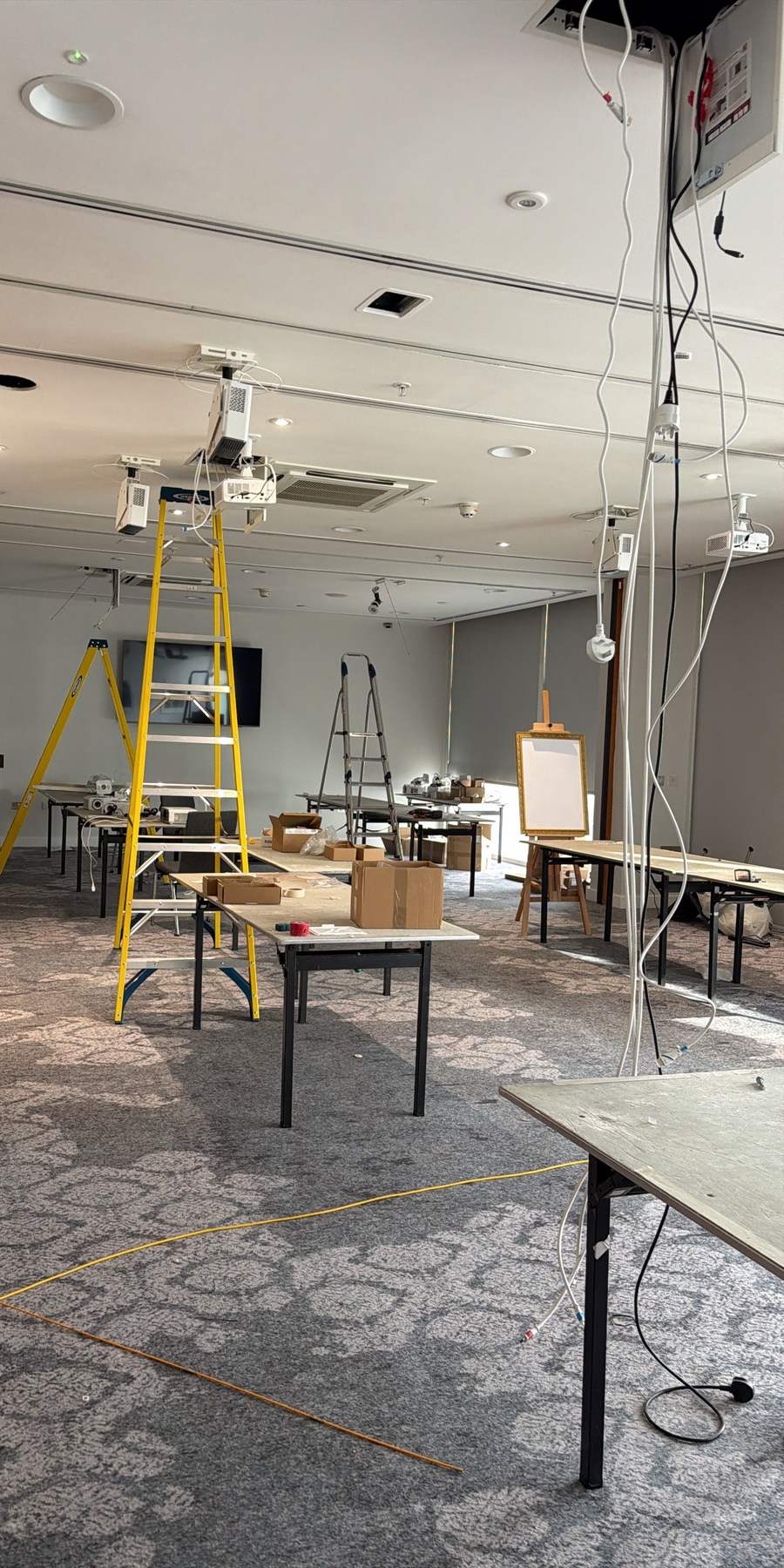 A function room on fit-out day: projectors hanging from open ceiling tiles on their cables, yellow ladders, boxes on the tables